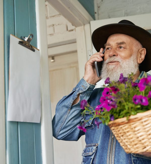 Low angle of senior man in cowboy hat speaking on cell phone and carrying basket with purple flowers